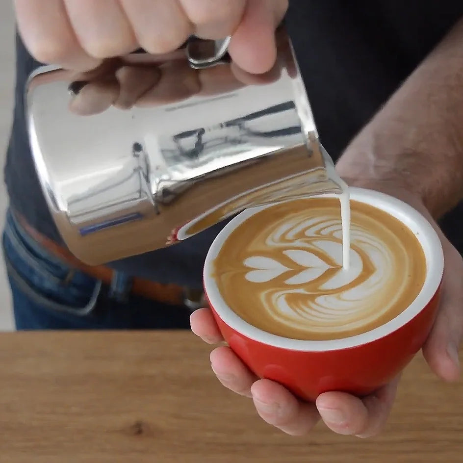 Person pouring milk into a red cup of coffee with latte art.