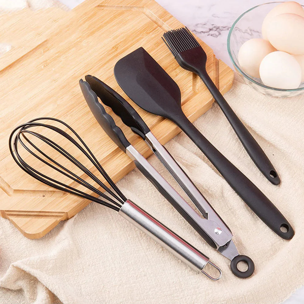 Set of black kitchen utensils including a whisk, tongs, spatula, and brush on a wooden cutting board with eggs in the background.