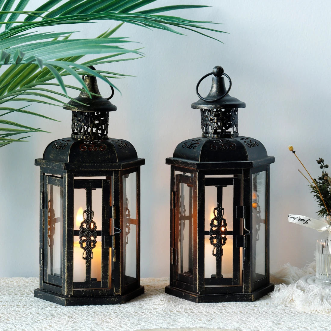 Two black lanterns with glass panels on a light surface with a plant in the background.