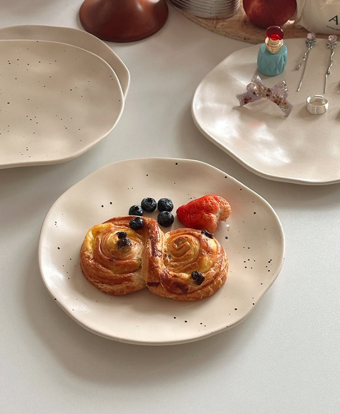 Two pastries with berries on a speckled ceramic plate, surrounded by other plates and items on a light surface.