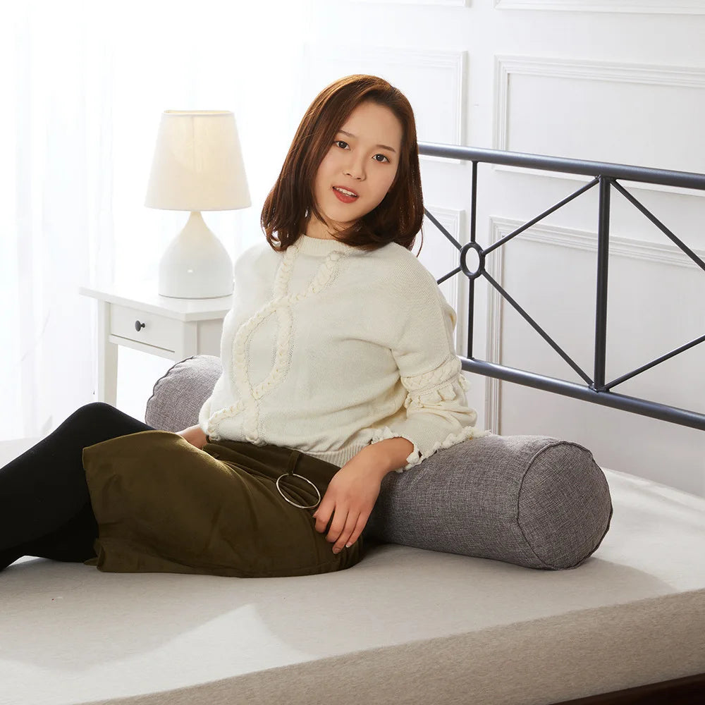 Woman sitting on a bed with a cylindrical pillow behind her back, in a bright room.