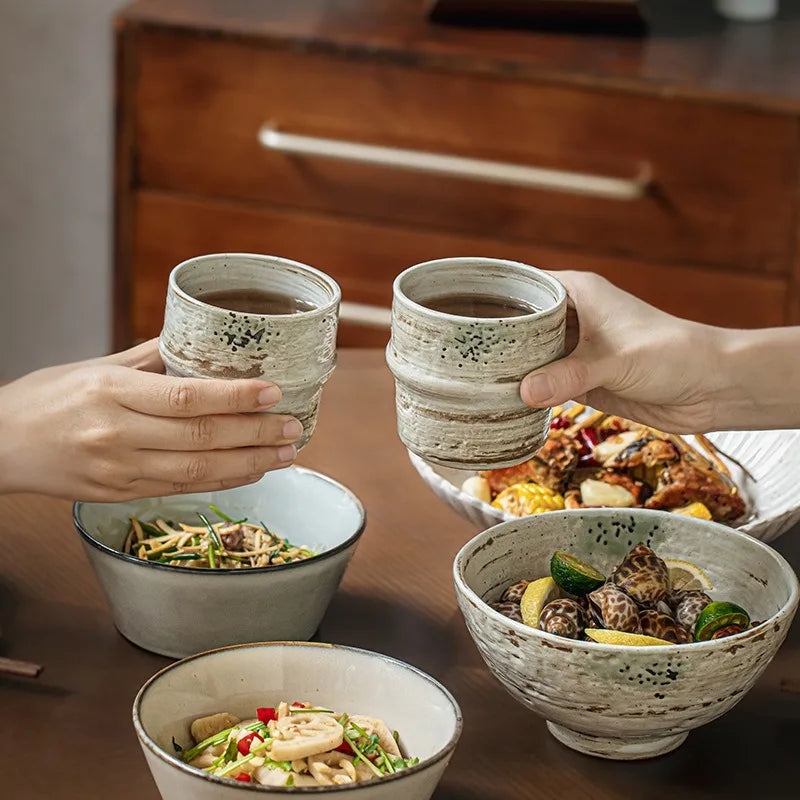 Two hands holding ceramic cups with a meal in bowls on a wooden table.