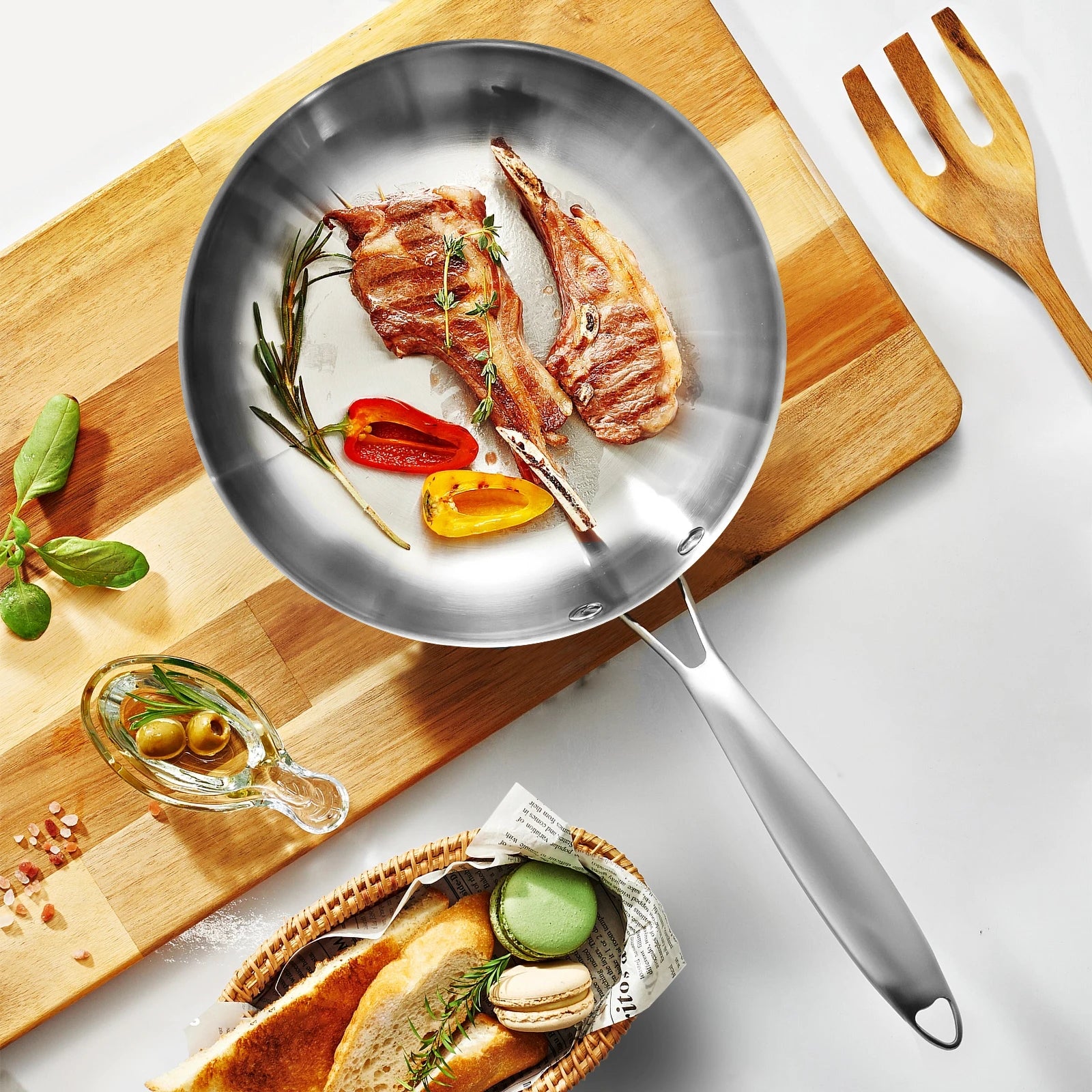 Stainless steel frying pan with cooked meat and vegetables on a wooden cutting board with bread and herbs.