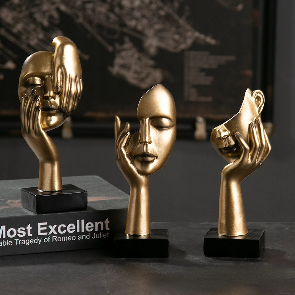 Three gold face sculptures with different poses on black bases with a blurred book in the background