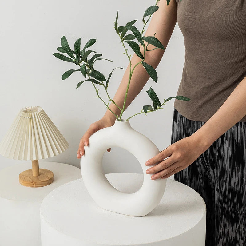Person holding a white abstract vase with green branches, with a small lamp in the background.