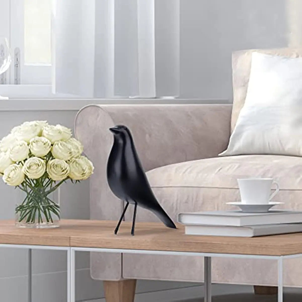 Black bird sculpture on a wooden table with a vase of white roses and a cup in a modern living room.