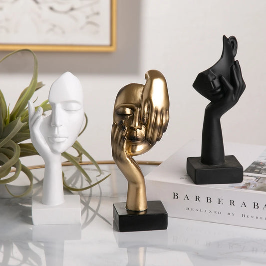 Three abstract sculpture face figurines with different poses on a surface with a book titled 'Barbera' in the background.