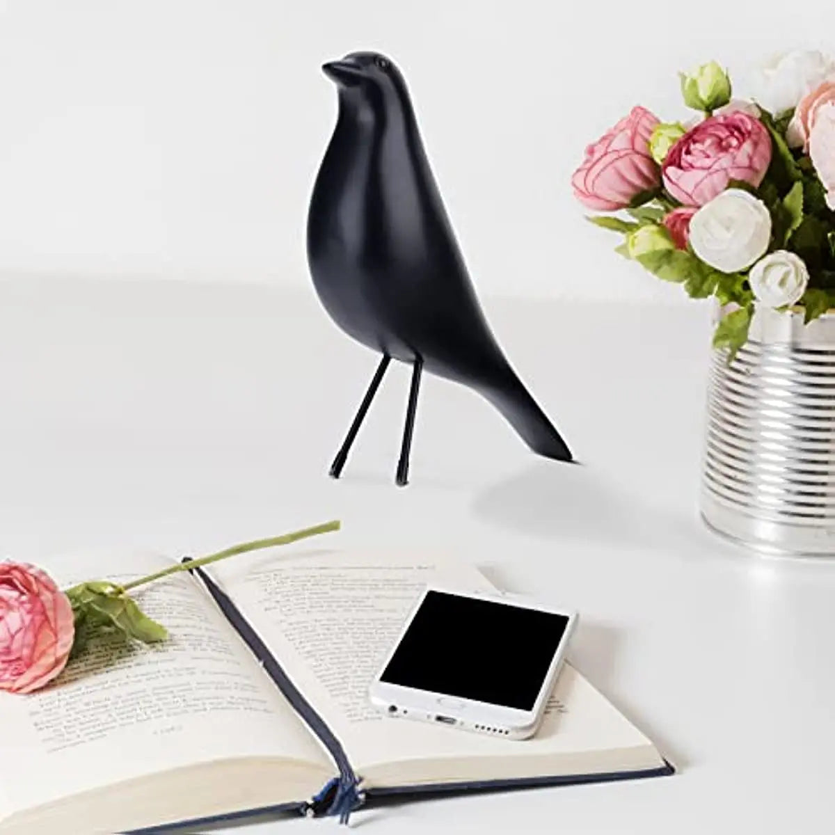 Black bird-shaped lamp on a table with an open book and flowers.