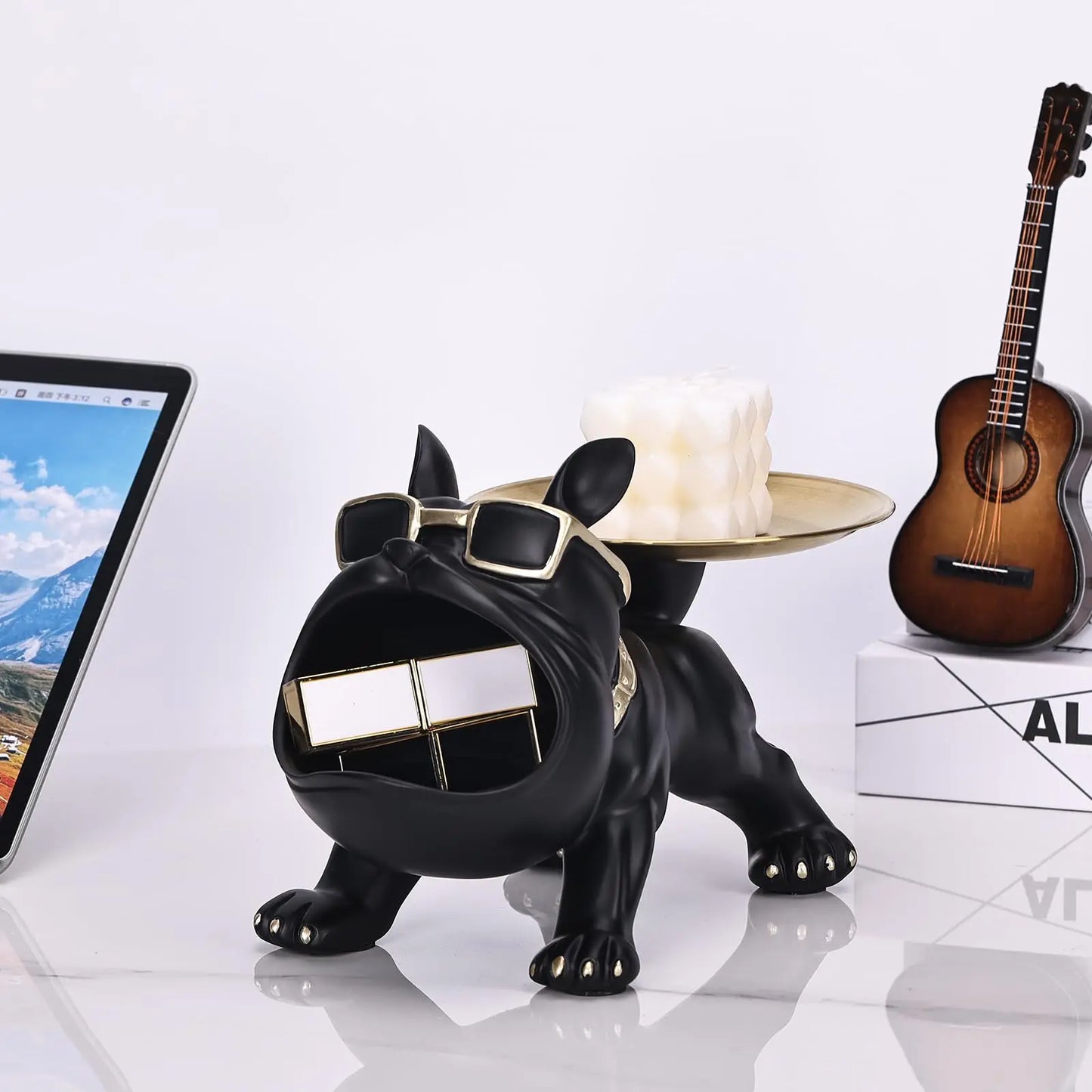 Black dog-shaped phone holder with sunglasses on a white surface with a tablet, candle, and guitar in the background.