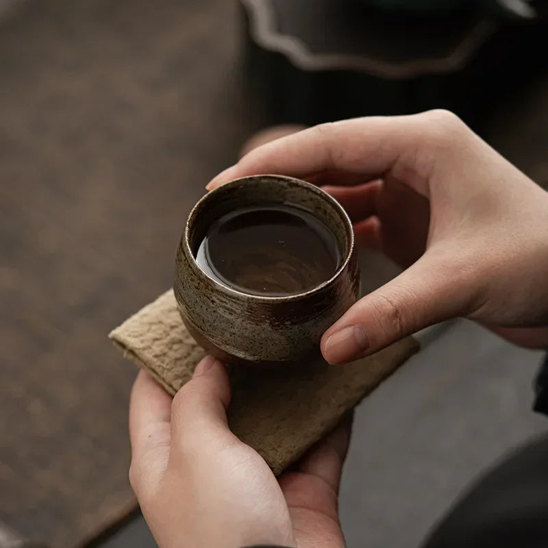 Person holding a small ceramic cup with a dark liquid on a wooden coaster.