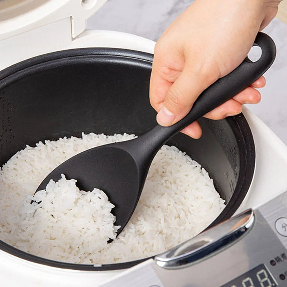 Black spoon scooping rice from a pot with a rice cooker in the background