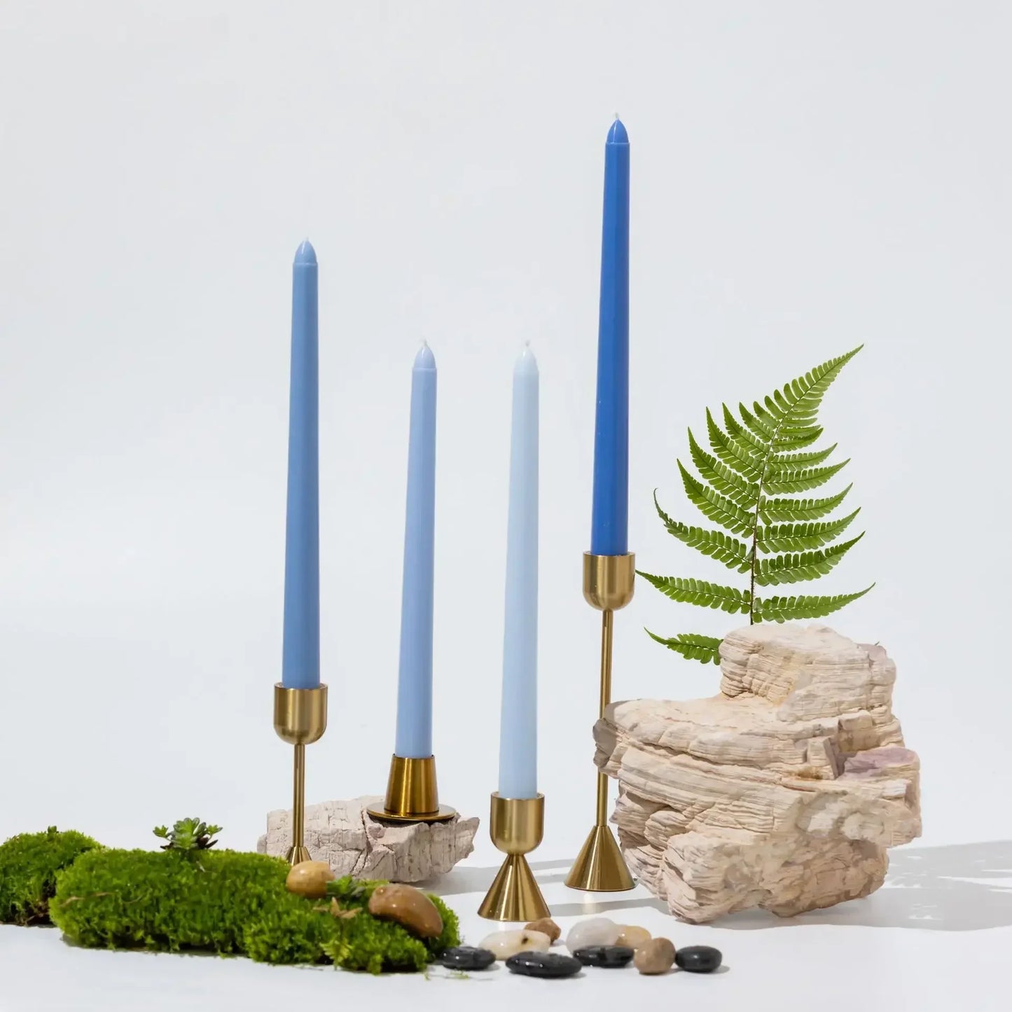 Decorative setup with blue candles in gold holders, rocks, and a fern on a white background